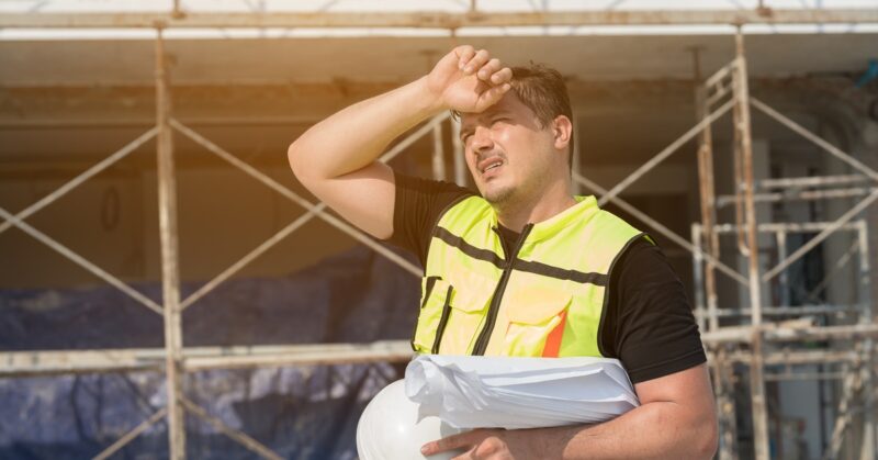 Construction worker in a neon safety vest shields his eyes from the sun while holding blueprints and a white hard hat on a scaffolding site.