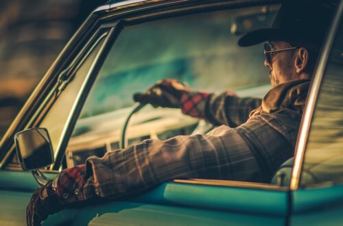 A man drives a vintage car at sunset, wearing a hat and gloves. His arm rests out the window, lit by warm lighting.