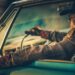 A man drives a vintage car at sunset, wearing a hat and gloves. His arm rests out the window, lit by warm lighting.
