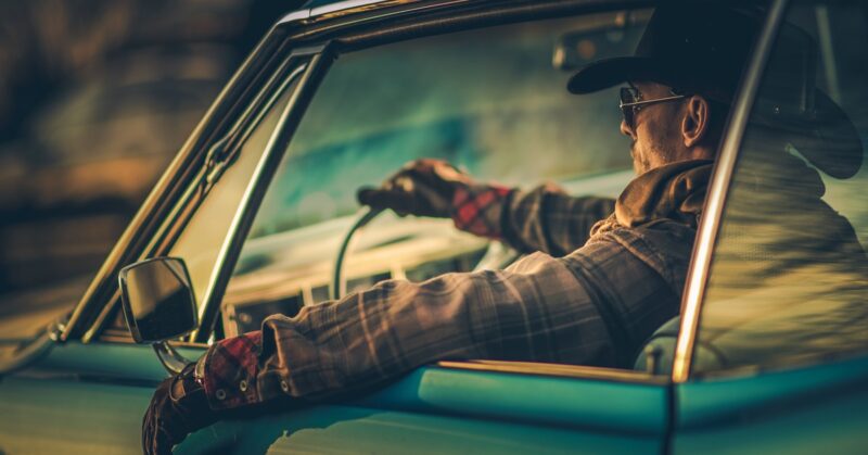 A man drives a vintage car at sunset, wearing a hat and gloves. His arm rests out the window, lit by warm lighting.