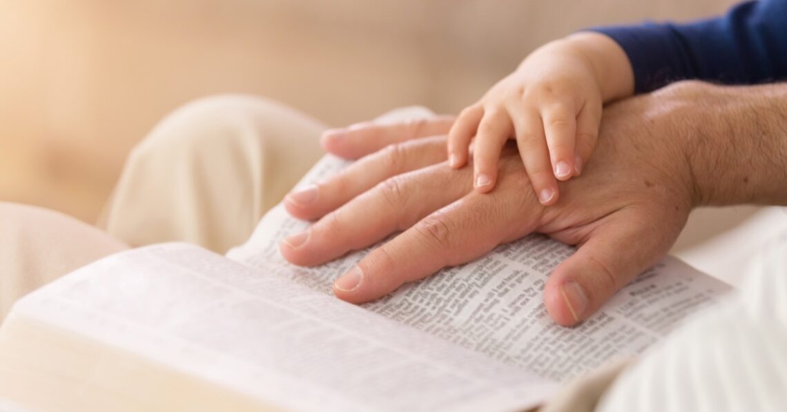 A close-up of a parent's hand on their Bible with their small child's hand on the adults. They're reading together.