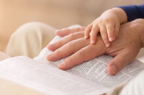 A close-up of a parent's hand on their Bible with their small child's hand on the adults. They're reading together.