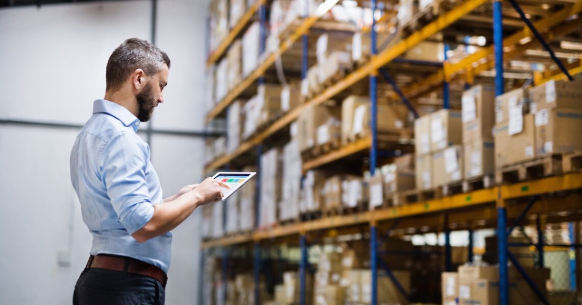Man in a light blue shirt uses a tablet to inspect inventory in a warehouse with tall orange shelves and stacked boxes.