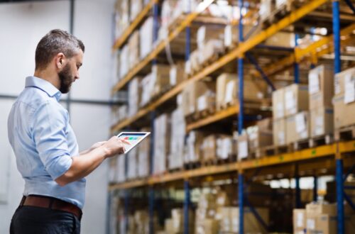 Man in a light blue shirt uses a tablet to inspect inventory in a warehouse with tall orange shelves and stacked boxes.