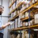 Man in a light blue shirt uses a tablet to inspect inventory in a warehouse with tall orange shelves and stacked boxes.