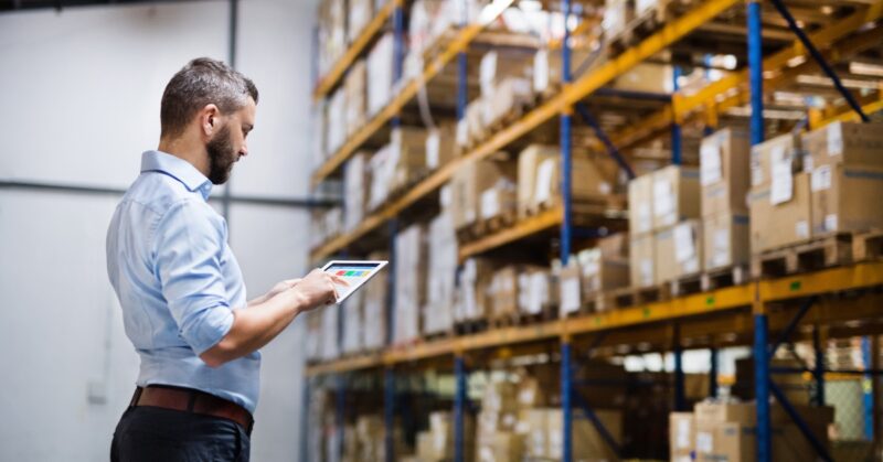 Man in a light blue shirt uses a tablet to inspect inventory in a warehouse with tall orange shelves and stacked boxes.