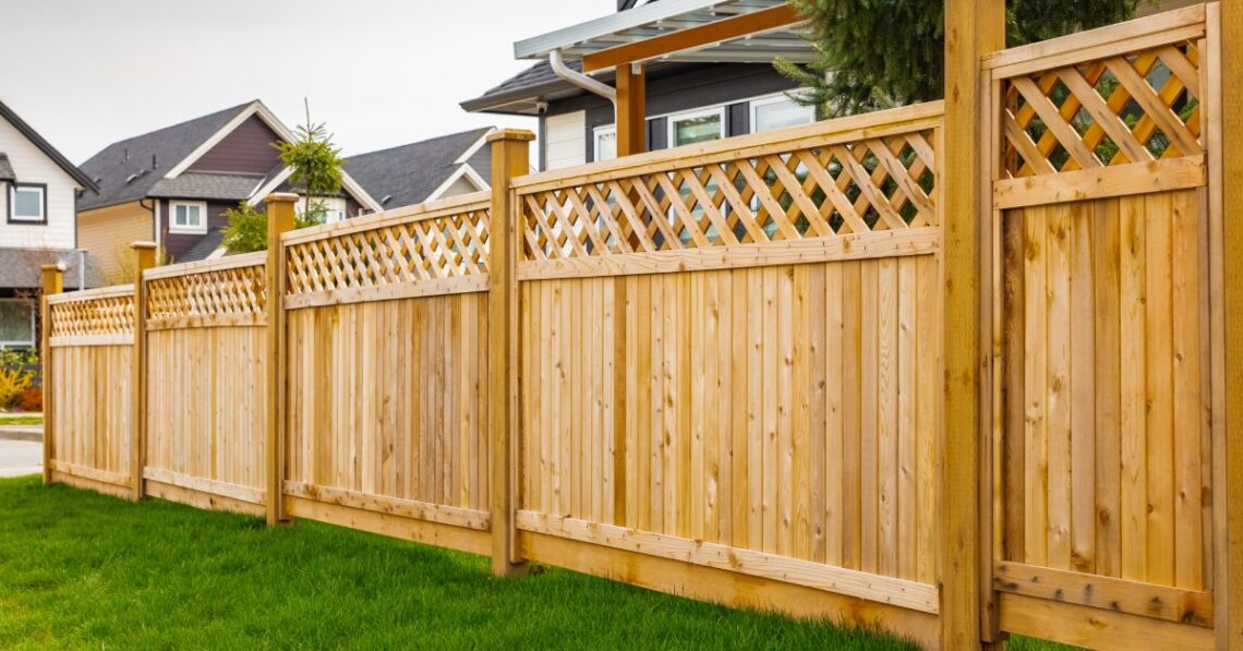 A long wooden privacy fence with lattice top runs along a green lawn in a suburban yard beside neighboring houses.
