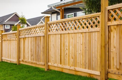 A long wooden privacy fence with lattice top runs along a green lawn in a suburban yard beside neighboring houses.