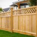 A long wooden privacy fence with lattice top runs along a green lawn in a suburban yard beside neighboring houses.
