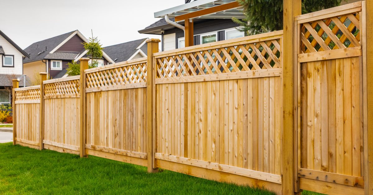 A long wooden privacy fence with lattice top runs along a green lawn in a suburban yard beside neighboring houses.