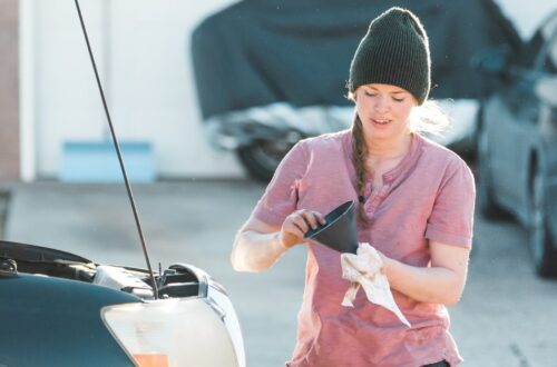 A woman in a beanie checks engine oil with a funnel and rag beside an open car hood in a bright outdoor setting.