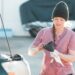 A woman in a beanie checks engine oil with a funnel and rag beside an open car hood in a bright outdoor setting.