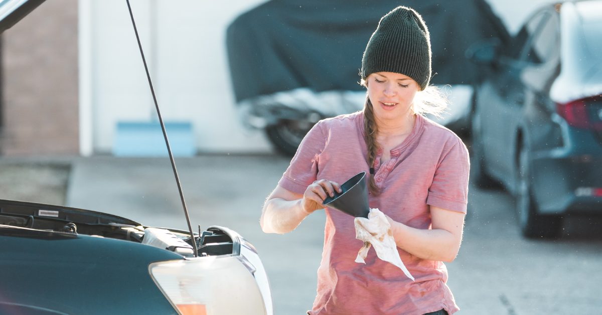 A woman in a beanie checks engine oil with a funnel and rag beside an open car hood in a bright outdoor setting.