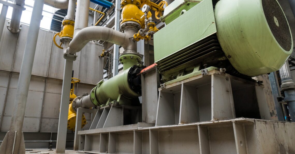 A large green machine sits on a raised platform, connected to multiple pipes and yellow valves in a warehouse.