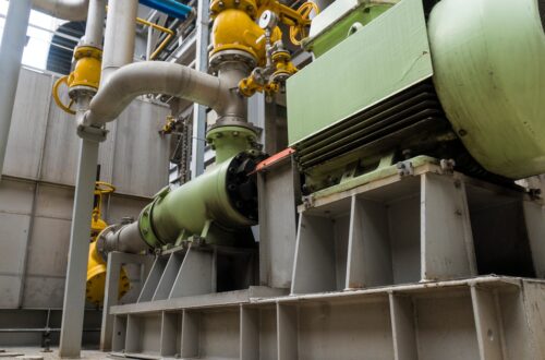 A large green machine sits on a raised platform, connected to multiple pipes and yellow valves in a warehouse.