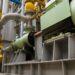 A large green machine sits on a raised platform, connected to multiple pipes and yellow valves in a warehouse.