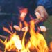 Four hands hold sticks with marshmallows over an open flame, roasting them above a fire pit with glowing flames.
