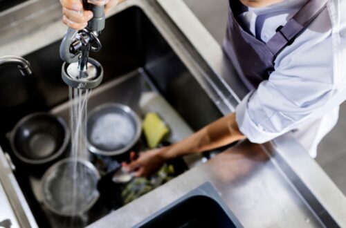 A restaurant worker is cleaning dishes in a large sink. They are spraying water on the dishes to start cleaning them.