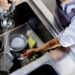 A restaurant worker is cleaning dishes in a large sink. They are spraying water on the dishes to start cleaning them.