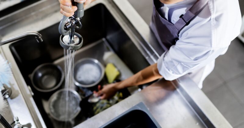 A restaurant worker is cleaning dishes in a large sink. They are spraying water on the dishes to start cleaning them.