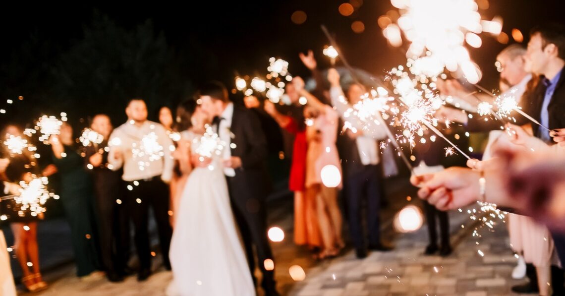 A blurry crowd of people around a happy married couple. Everyone is holding sparklers out while the couple kisses.