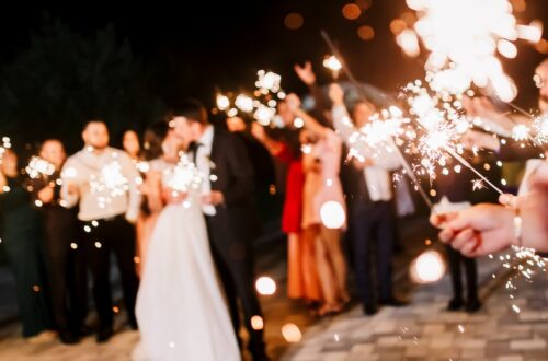 A blurry crowd of people around a happy married couple. Everyone is holding sparklers out while the couple kisses.