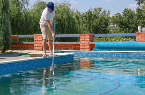 A man in a ballcap, t-shirt, and shorts using a long-handled vacuum with an attached tube to clean a swimming pool.
