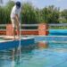 A man in a ballcap, t-shirt, and shorts using a long-handled vacuum with an attached tube to clean a swimming pool.