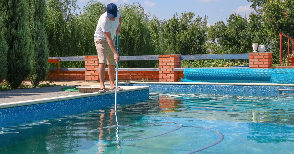 A man in a ballcap, t-shirt, and shorts using a long-handled vacuum with an attached tube to clean a swimming pool.