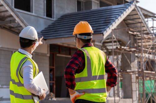 An engineer points out house construction details to a supervisor at a building site, both wearing hard hats.