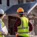 An engineer points out house construction details to a supervisor at a building site, both wearing hard hats.