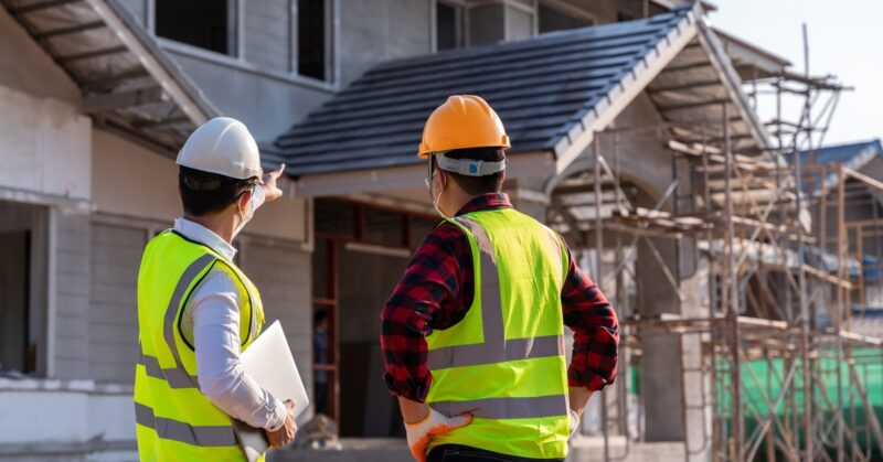 An engineer points out house construction details to a supervisor at a building site, both wearing hard hats.