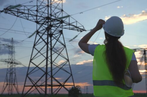 An electrician in a safety vest and a hard hat looking up at transmission towers. The sun is low in the distance.