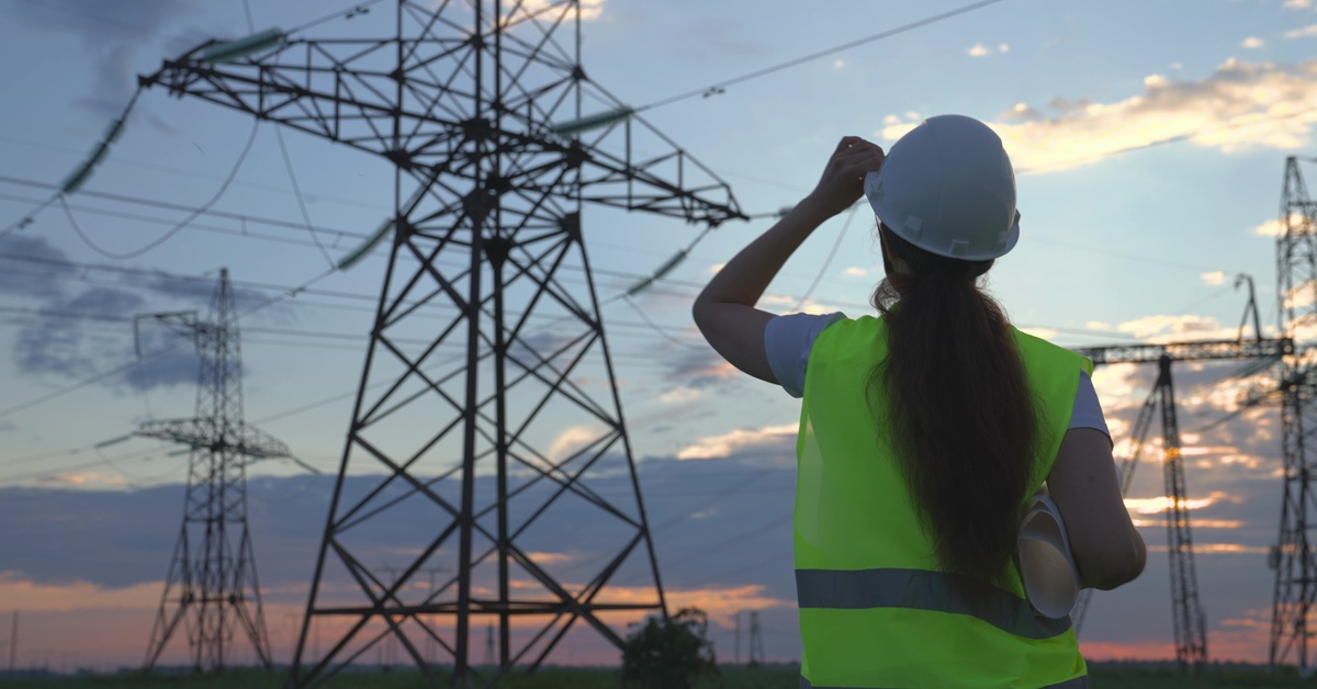 An electrician in a safety vest and a hard hat looking up at transmission towers. The sun is low in the distance.