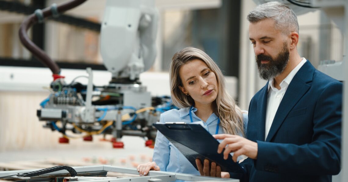 A businessman and engineer both looking at a clipboard the man is holding. There is a manufacturing robot behind them.