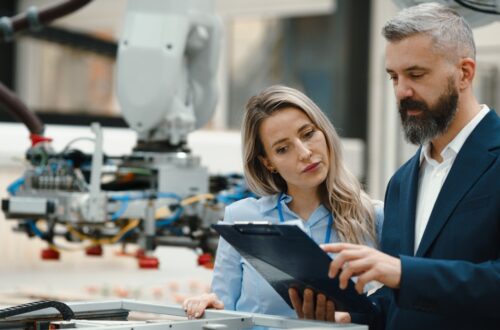 A businessman and engineer both looking at a clipboard the man is holding. There is a manufacturing robot behind them.