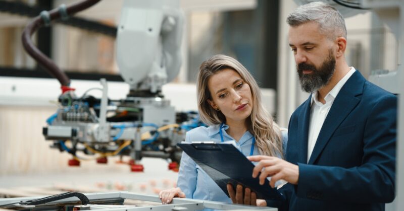 A businessman and engineer both looking at a clipboard the man is holding. There is a manufacturing robot behind them.