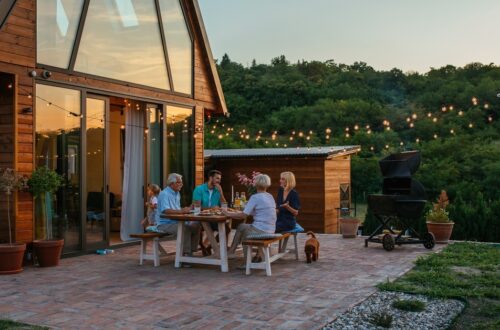 Family gathered around a wooden outdoor table on a brick patio at dusk, sharing a meal under string lights by a cabin-like home.