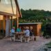 Family gathered around a wooden outdoor table on a brick patio at dusk, sharing a meal under string lights by a cabin-like home.