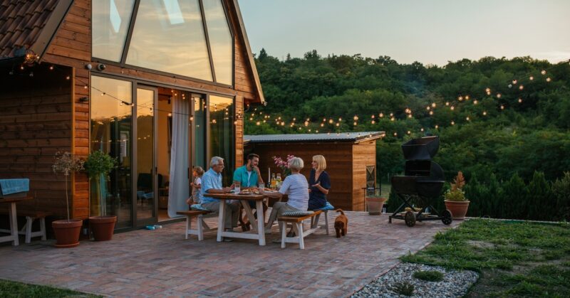 Family gathered around a wooden outdoor table on a brick patio at dusk, sharing a meal under string lights by a cabin-like home.