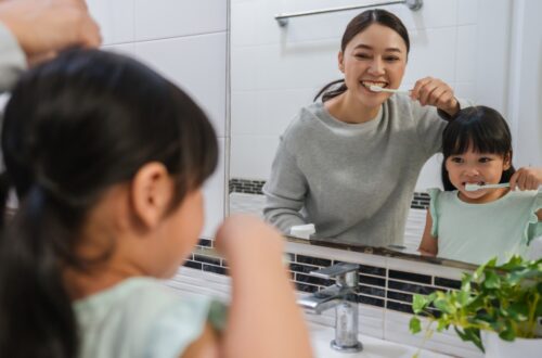A mother and daughter brushing their teeth as they look at themselves in the bathroom mirror above the sink.