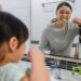 A mother and daughter brushing their teeth as they look at themselves in the bathroom mirror above the sink.