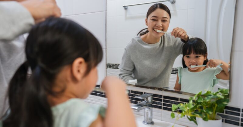 A mother and daughter brushing their teeth as they look at themselves in the bathroom mirror above the sink.