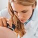 A dermatologist using a handheld device to closely examine the skin on another person's shoulder in a bright room.