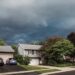 Suburban homes under dark, looming storm clouds, with trees swaying and a quiet street signaling an approaching severe storm.