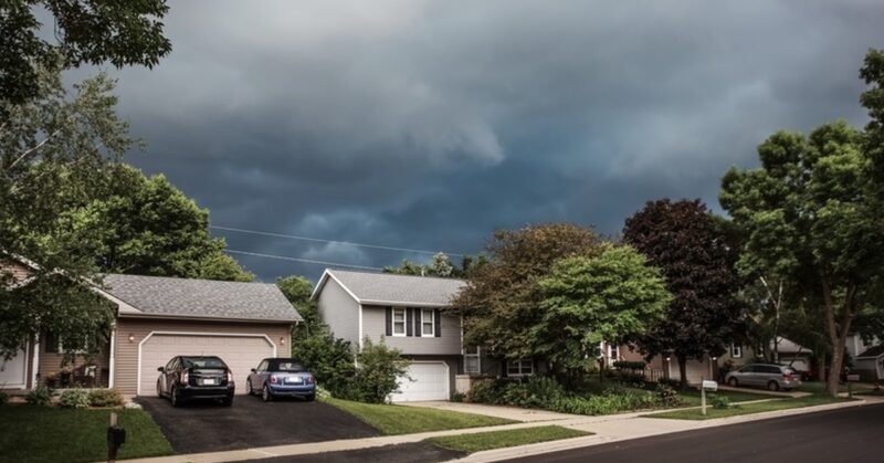 Suburban homes under dark, looming storm clouds, with trees swaying and a quiet street signaling an approaching severe storm.