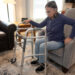 An older woman sits on the edge of a grey chair in her living room. She has a walker in front of her with one arm on it.