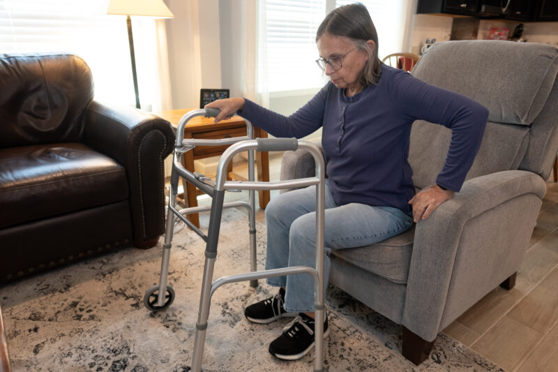 An older woman sits on the edge of a grey chair in her living room. She has a walker in front of her with one arm on it.