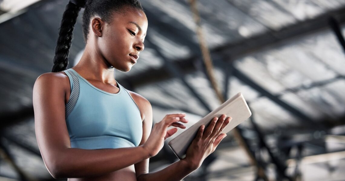 A low-angle view of a young Black woman in a light-blue athletic outfit using a tablet at a facility.