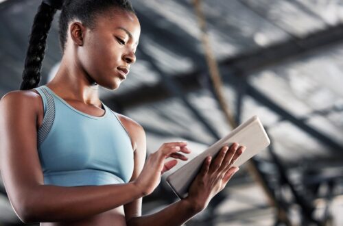 A low-angle view of a young Black woman in a light-blue athletic outfit using a tablet at a facility.
